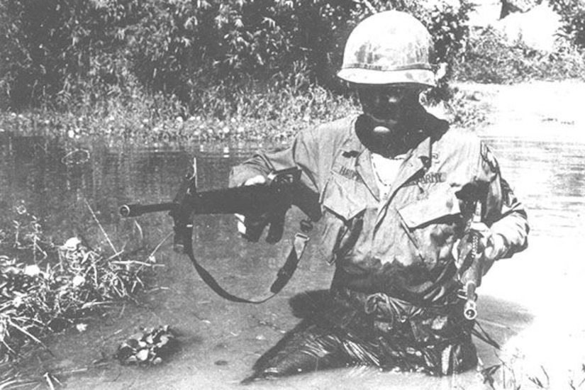 A soldier wearing battle fatigues and a helmet wades through waist-deep water in a Vietnam swamp while carrying a rifle.
