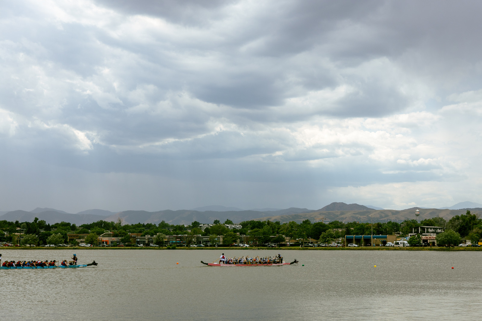 Two long boats each rowed by dozens of people, a red one in the middle of the frame and a green one truncated on the left, two floats on the right, and rain clouds over the mountains in the background