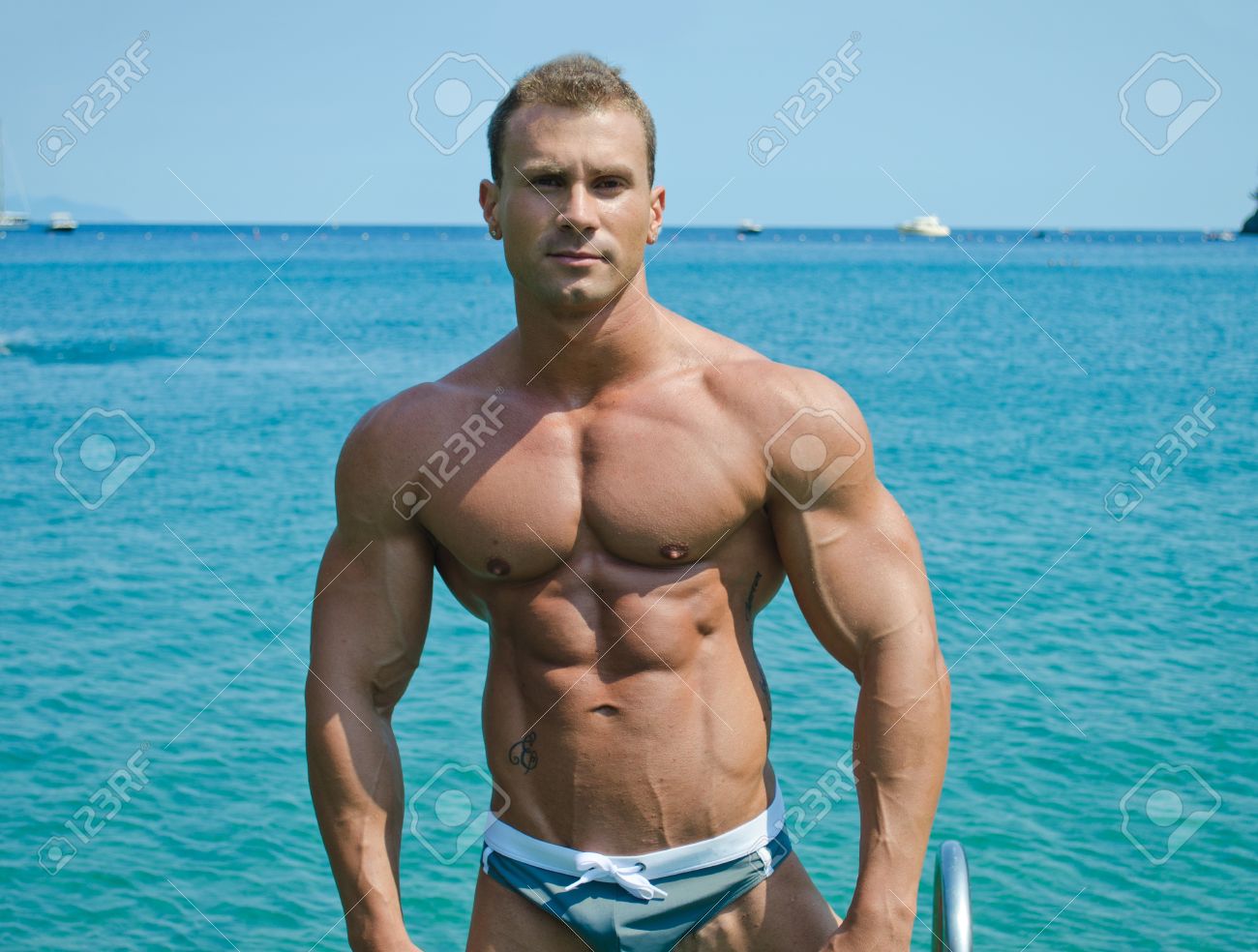 20680011-Handsome-young-bodybuilder-standing-with-sea-or-ocean-behind-showing-muscular-torso-pecs-arms-and-ab-Stock-Photo.jpg