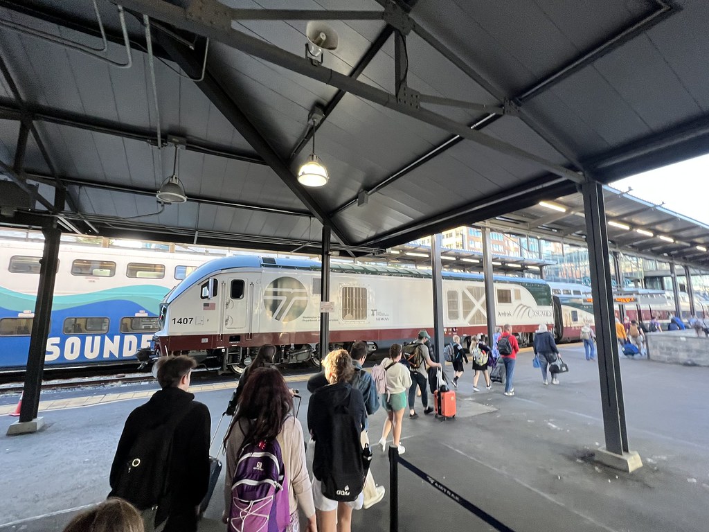 A file of people with backpacks and luggage walking towards a Siemens Charger locomotive and train cars