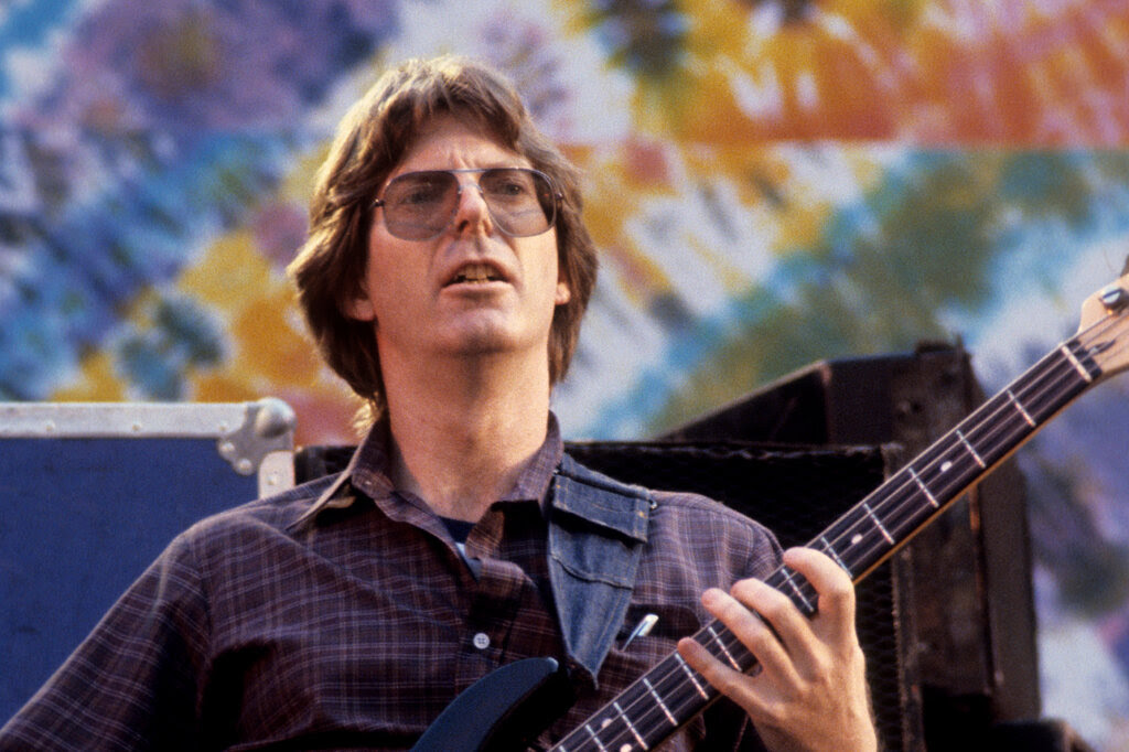 A portrait of Phil Lesh holding his bass guitar and standing against a colorful tie-dye background.