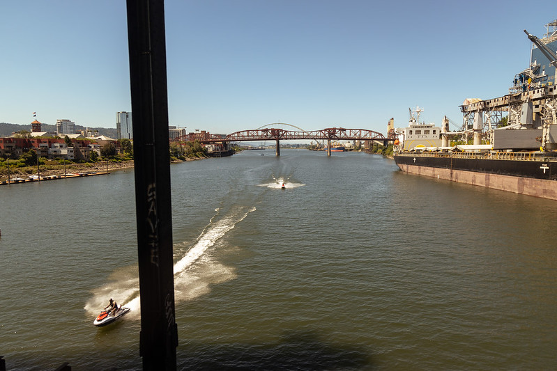 Two jet skis speeding down a river, with a bridge and buildings in the background, seen from above